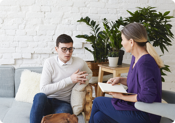 A man in glasses talks to a woman holding a notebook. They sit on a sofa in a bright room with plants.