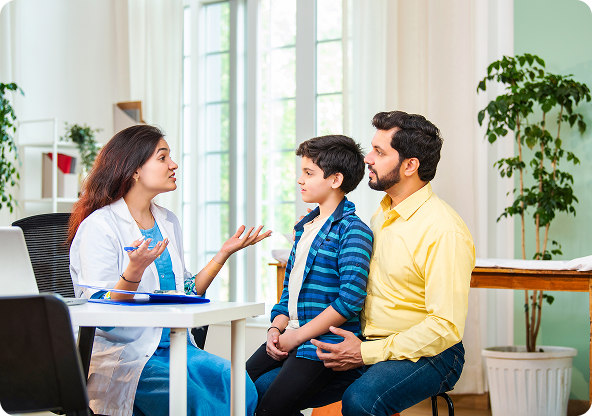 A doctor consults with a boy and his father in an office. The doctor gestures with her hands while talking, and the father has his arm around the boy.