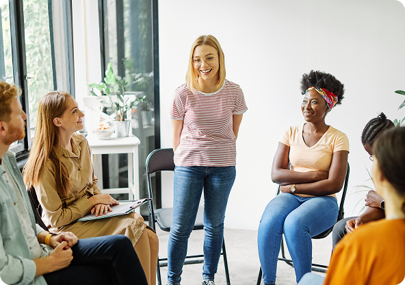 A group therapy session. A woman smiles while standing, surrounded by people seated in chairs.