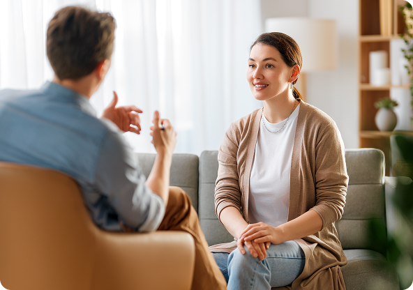 Man and woman in a counseling session. The woman smiles at the man, who gestures with his hands. They're seated in a bright room.