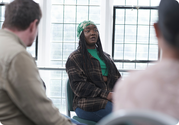 A Black woman with long braids wearing a green top and plaid shirt speaks in a group therapy setting.