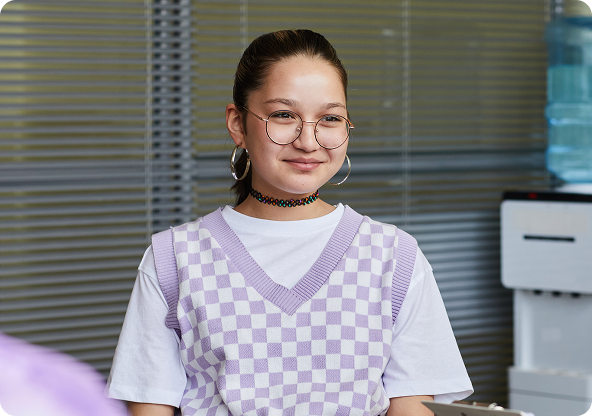 A young woman with glasses smiles, wearing a purple checkered vest and a choker necklace, standing near a water cooler.
