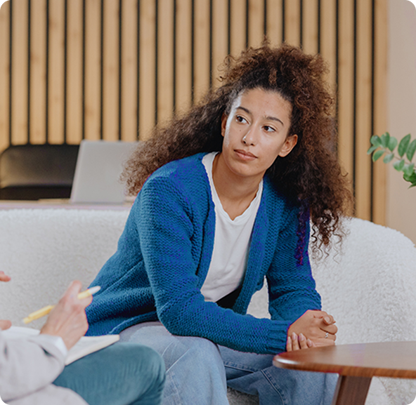 A young woman with curly hair sits on a couch, looking thoughtful, while a person holding a notepad is next to her.