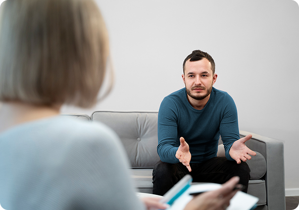 Man sitting on a gray couch talking to a therapist in an office setting; the therapist is holding notes.