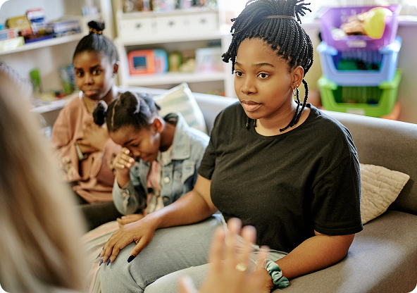 A woman with braids talks, sitting on a couch with two girls. One girl looks sad, the other watches. The setting appears to be indoors.