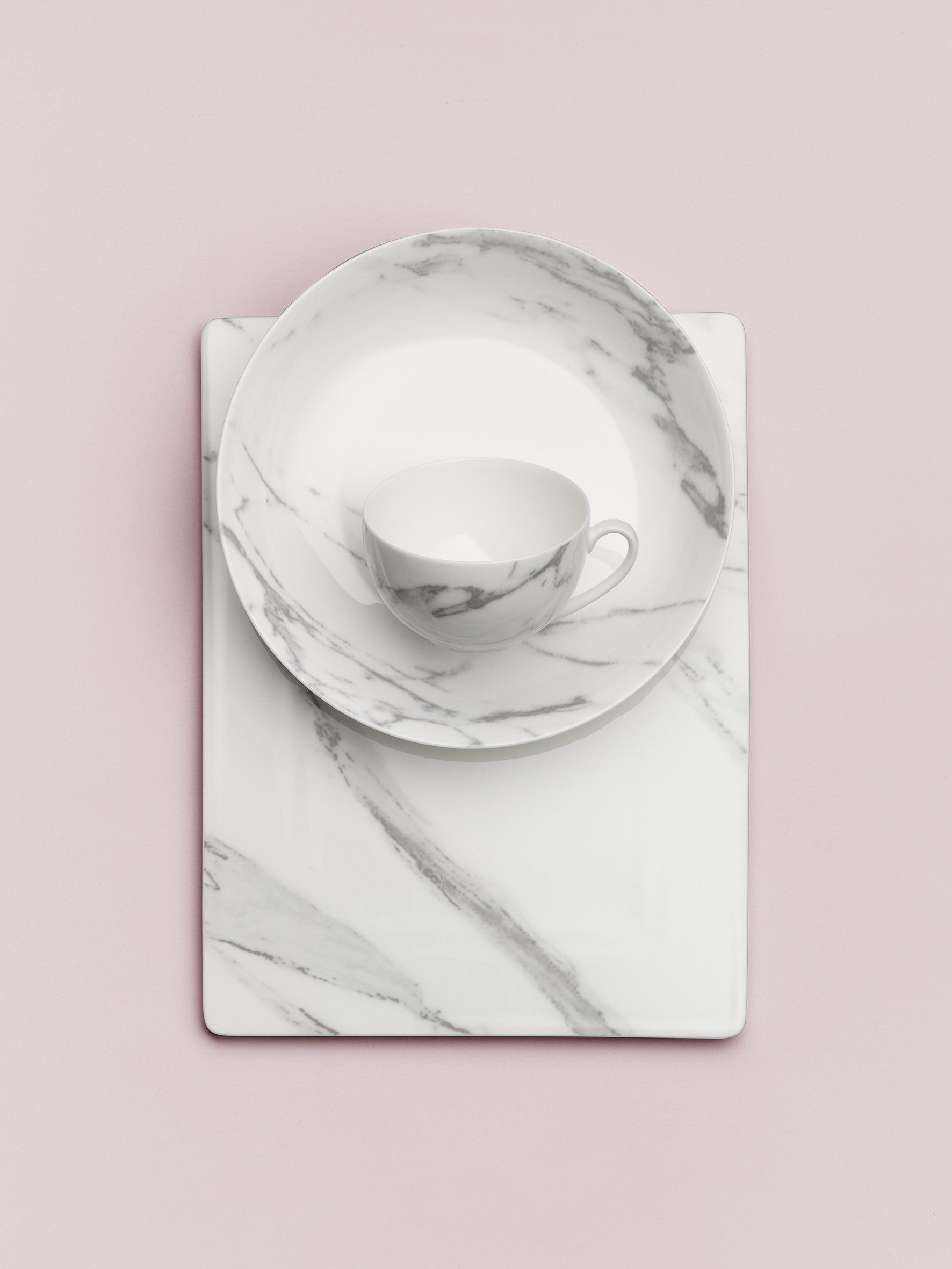 Ein Teller, eine Untertasse und eine Tasse auf einem Marmortablett auf rosa Hintergrund.
