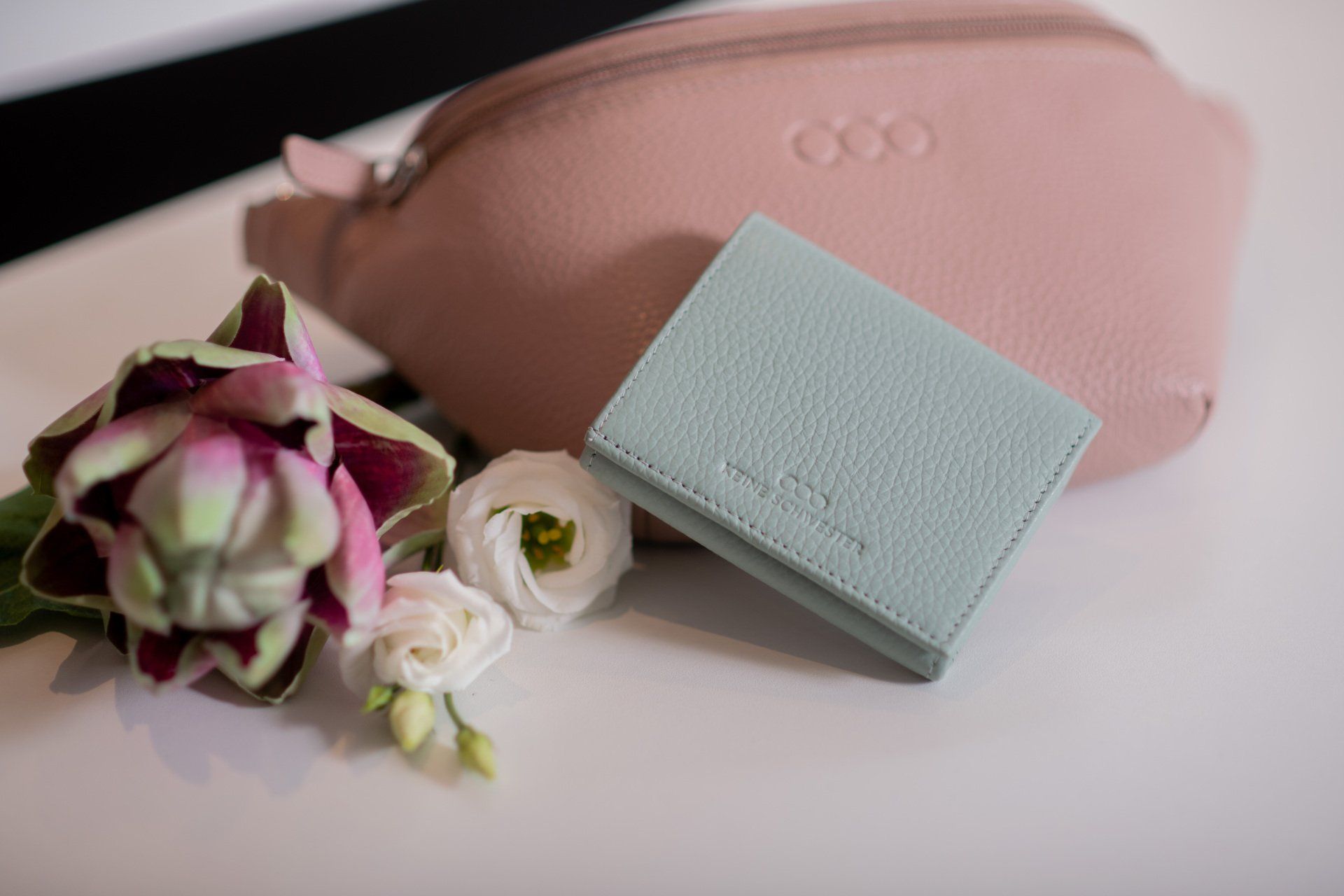 Eine rosa Handtasche und eine blaue Brieftasche liegen auf einem Tisch neben Blumen.