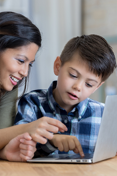 Woman and young boy using a laptop, woman pointing, boy looking intently, both smiling, indoors.