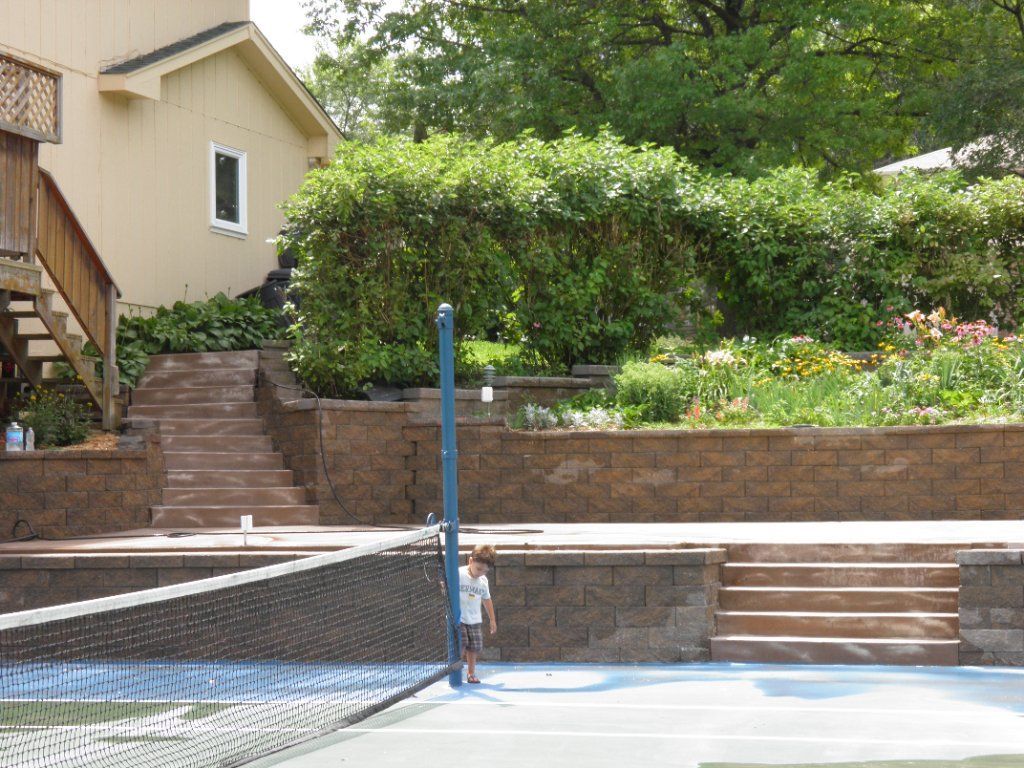 Tennis court with net, steps, and a person near the net, with a terraced garden and building in the background. Tennis court with net, steps, and a person near the net, with a terraced garden and building in the background.