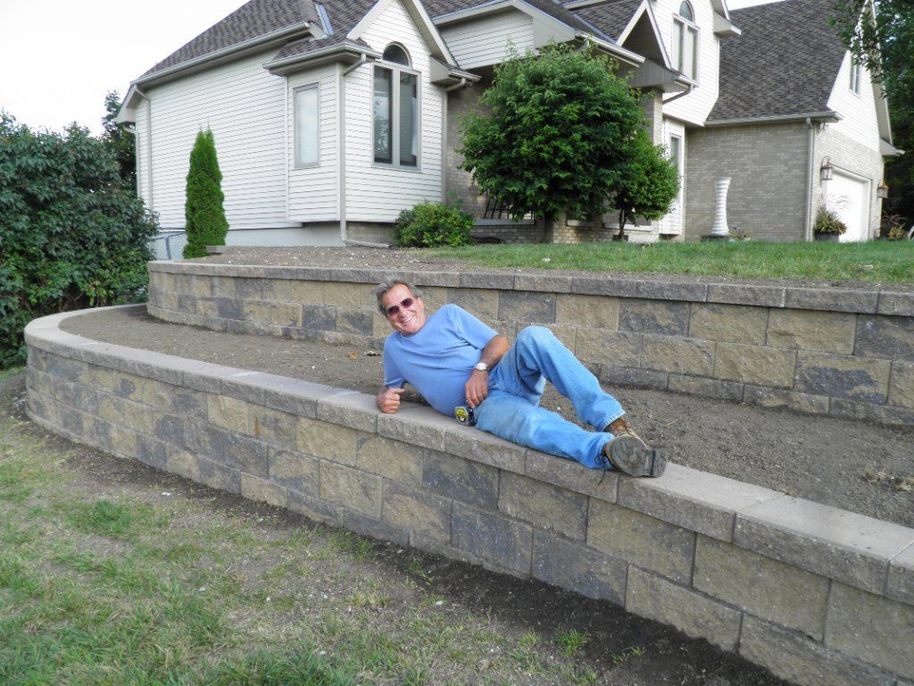 Man reclining on a stone retaining wall in front of a house. He’s wearing blue jeans and a light blue shirt. Man reclining on a stone retaining wall in front of a house. He’s wearing blue jeans and a light blue shirt.