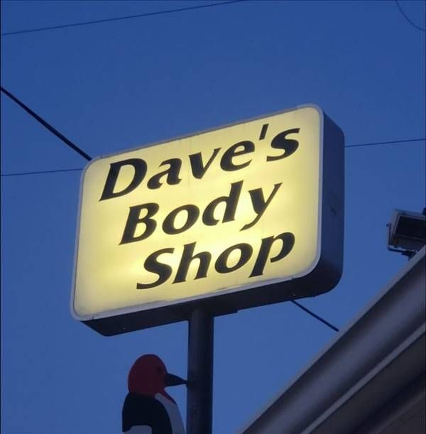 Sign for Dave's Body Shop; illuminated rectangular sign with black text on a yellow background, mounted outdoors at dusk.