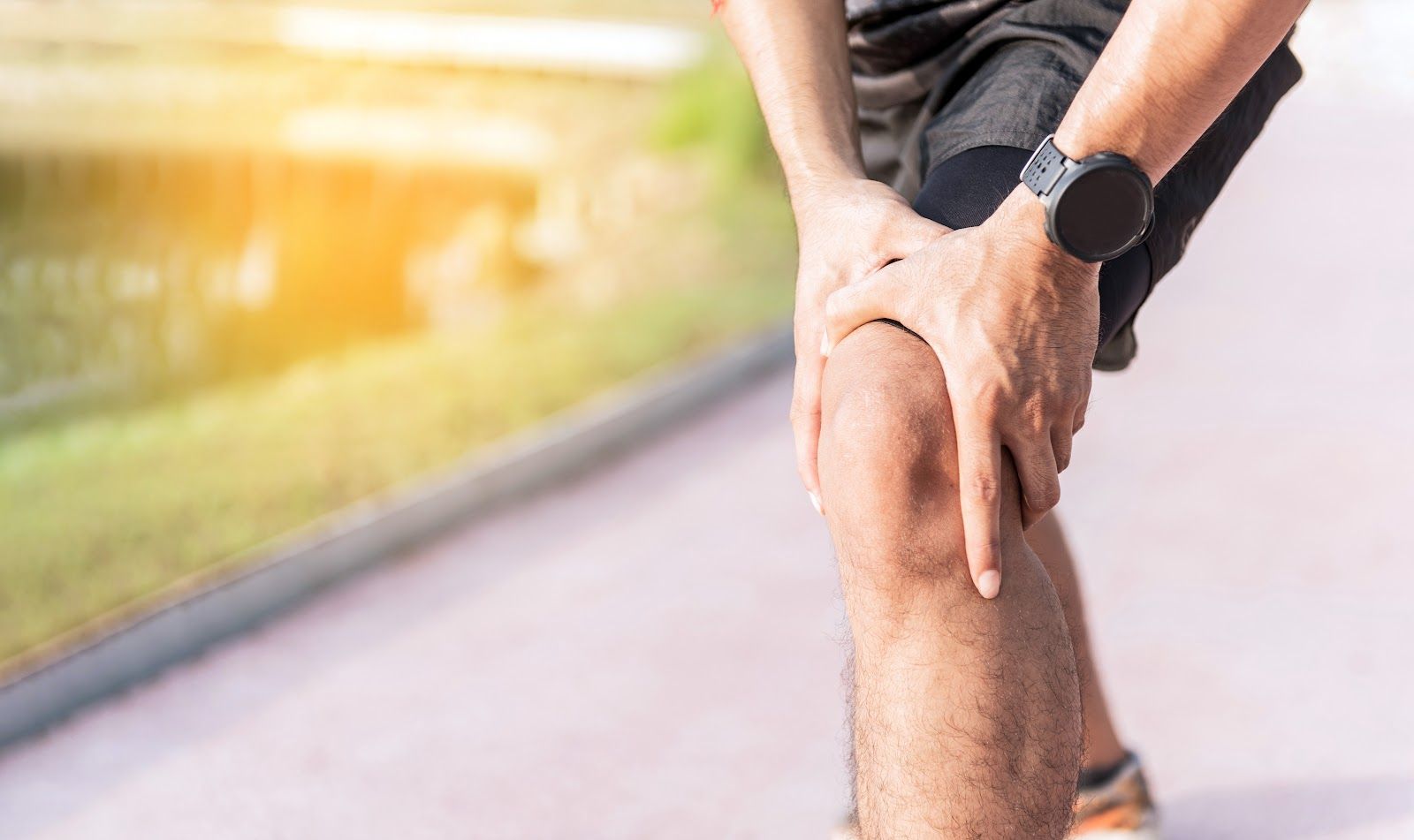 Runner clutching knee in pain on a blurred outdoor path, wearing a black smartwatch.