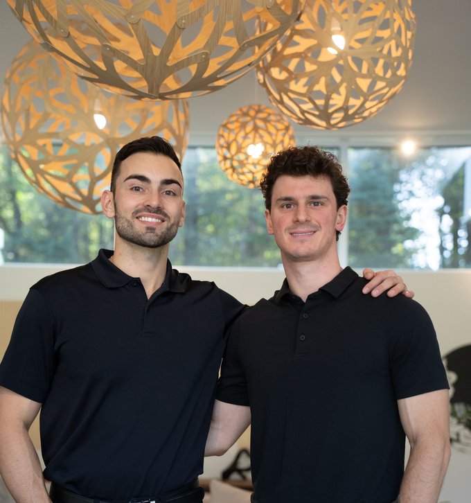 Two smiling men in black polos posing against a wood-paneled wall.