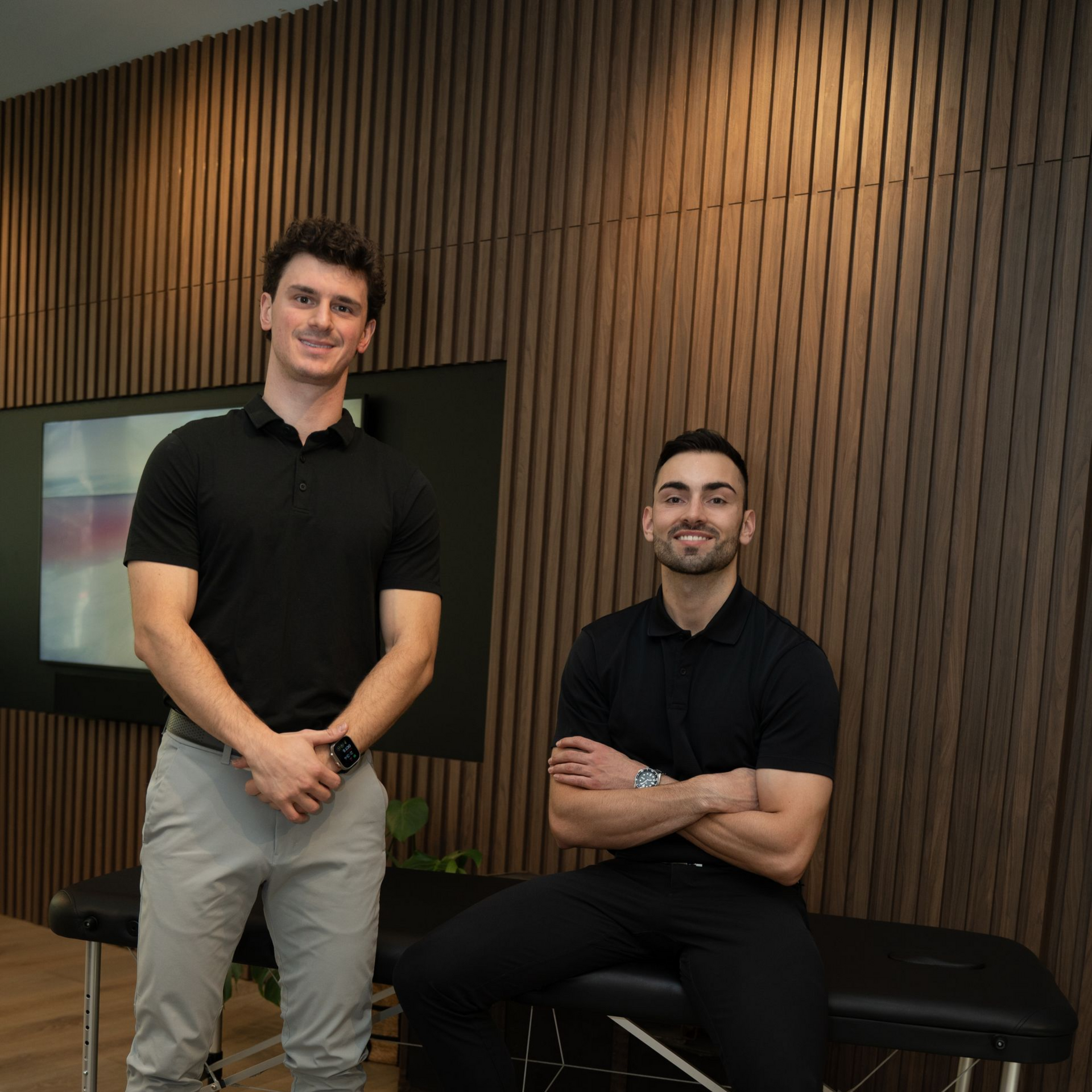 Two people in black polo shirts posing by a bench in a modern wood-paneled room