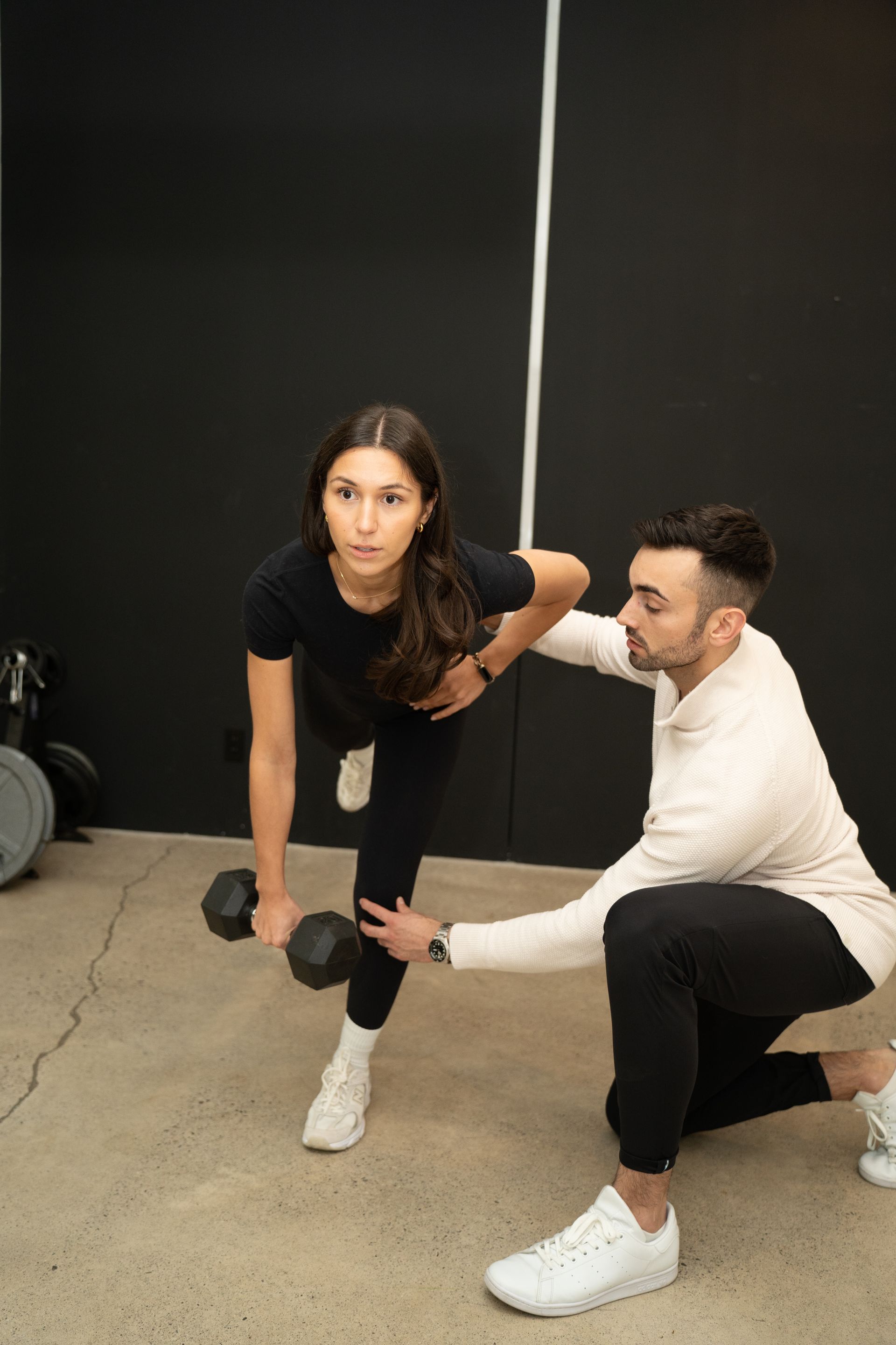 Trainer assisting client with dumbbell exercise in a gym, both crouched on a concrete floor.