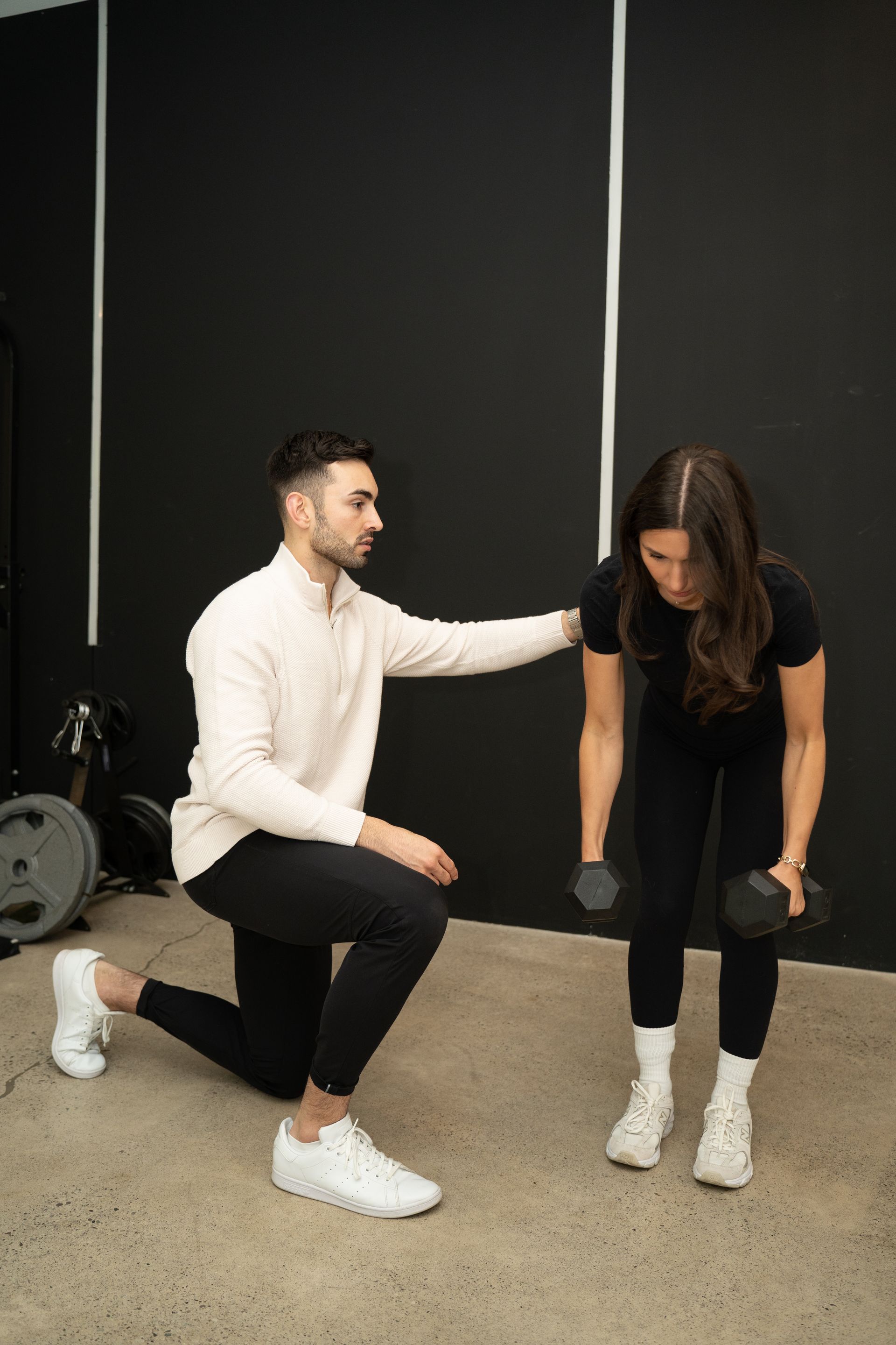 Trainer kneels beside a woman in a gym, coaching her during a dumbbell exercise.