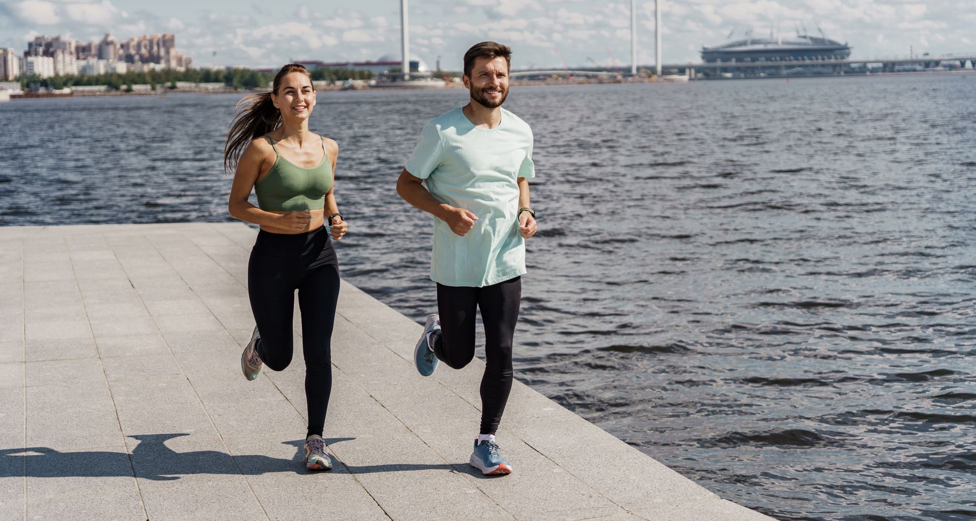 Two people running along a waterfront promenade, with water and distant buildings behind them.