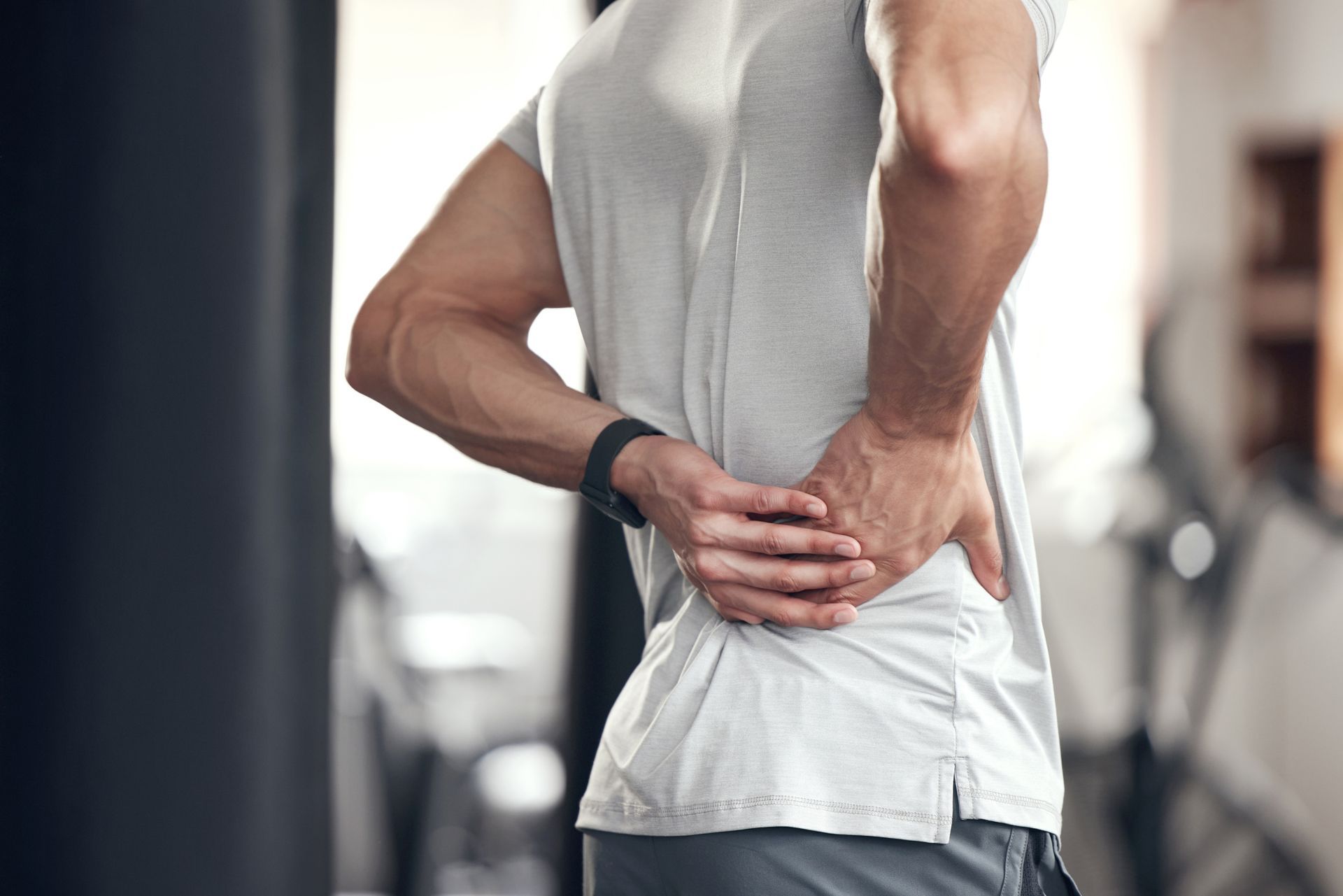 Person in a white shirt holding their lower back, standing indoors in a gym-like setting.