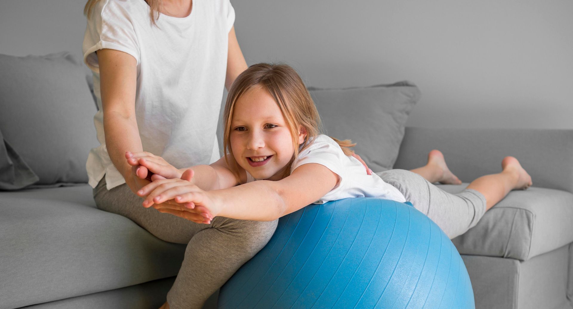 Child smiling on a gray sofa while doing physical therapy with a blue exercise ball and adult guidance