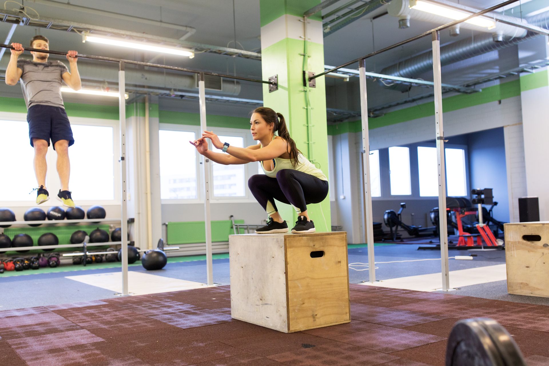 Two people exercising in a gym, one jumping to a box and one lifting with arms extended