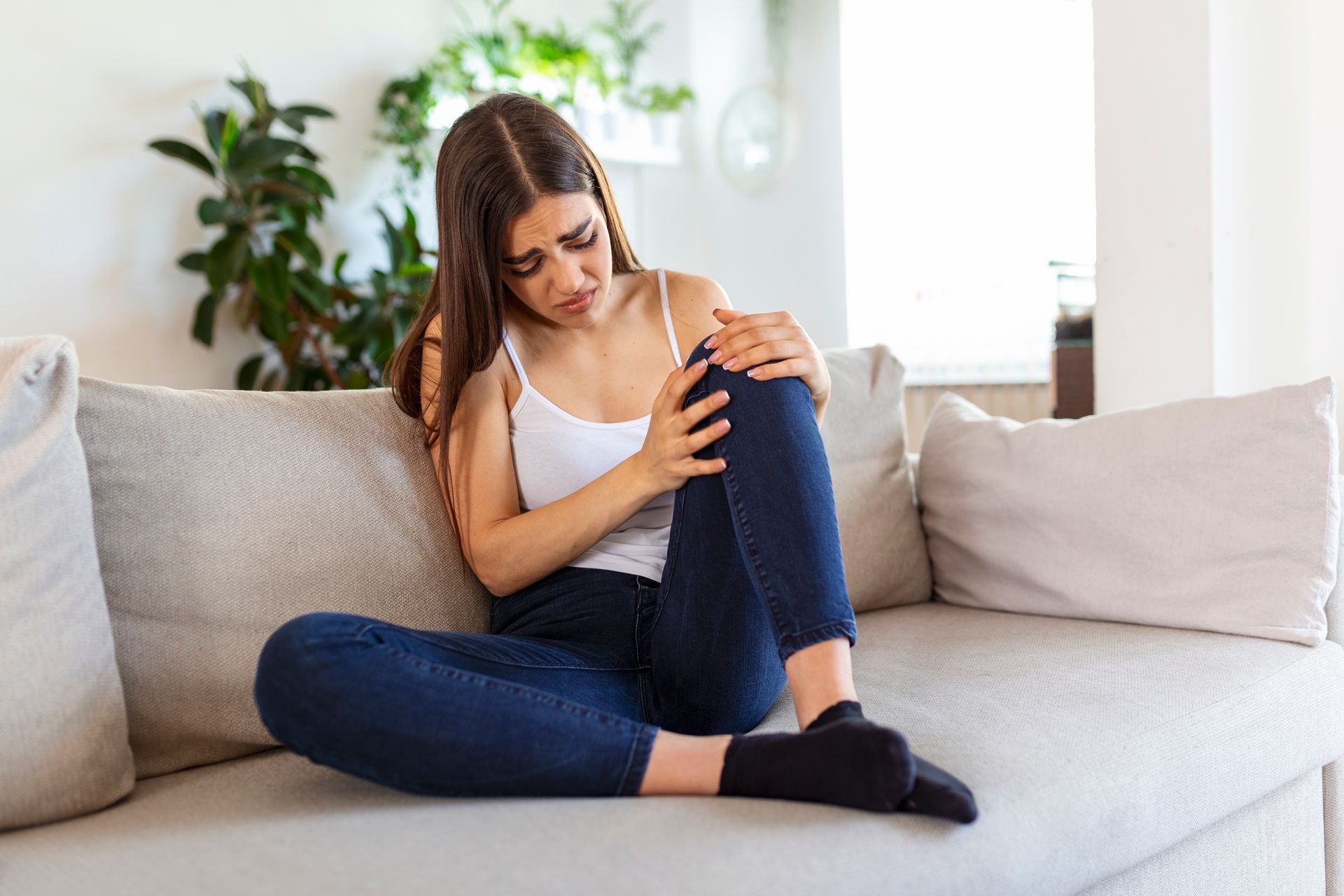 Woman sitting on a couch, holding her knee in discomfort indoors.