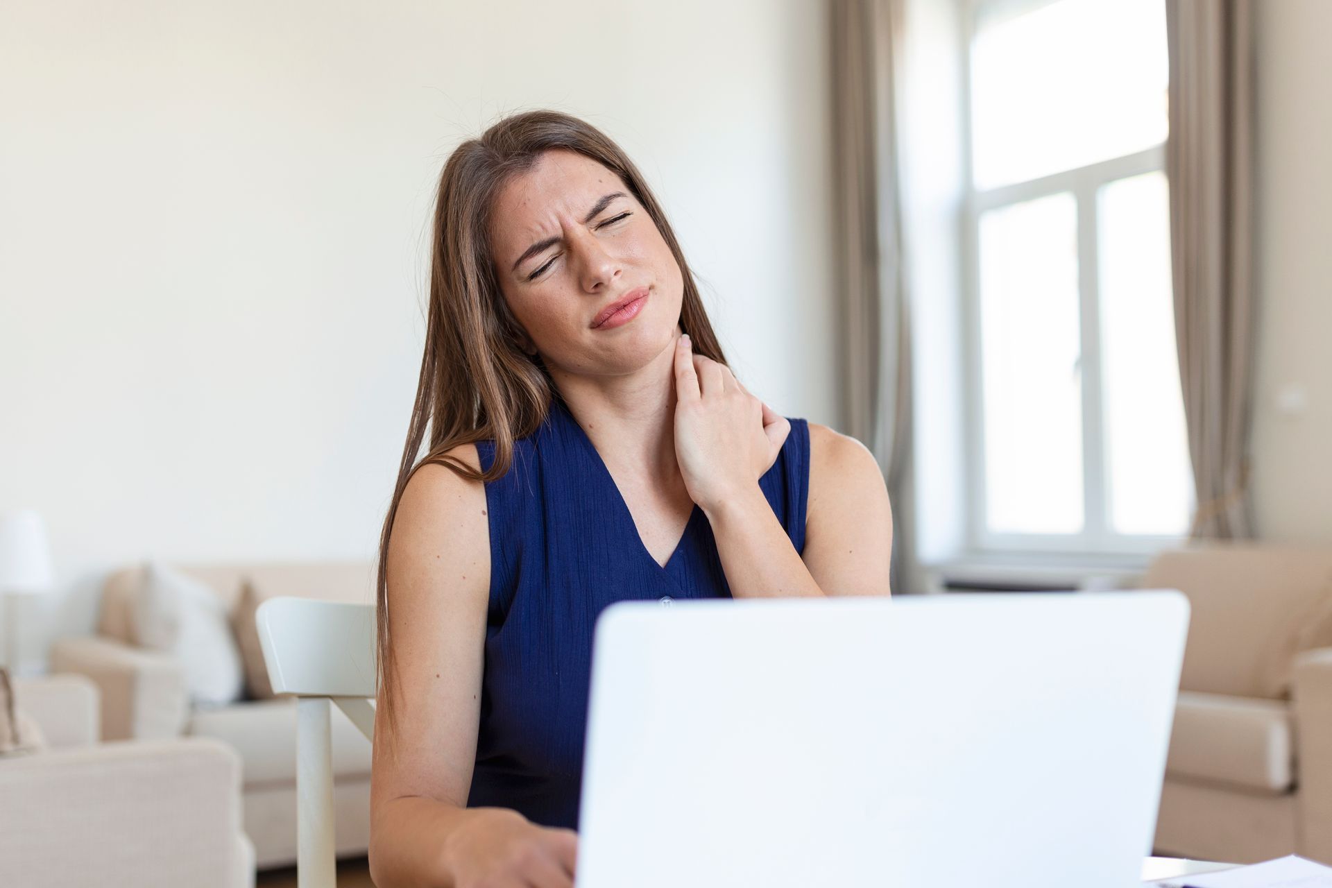Woman in a blue top rubbing her neck while sitting behind a laptop, appearing to feel neck pain.
