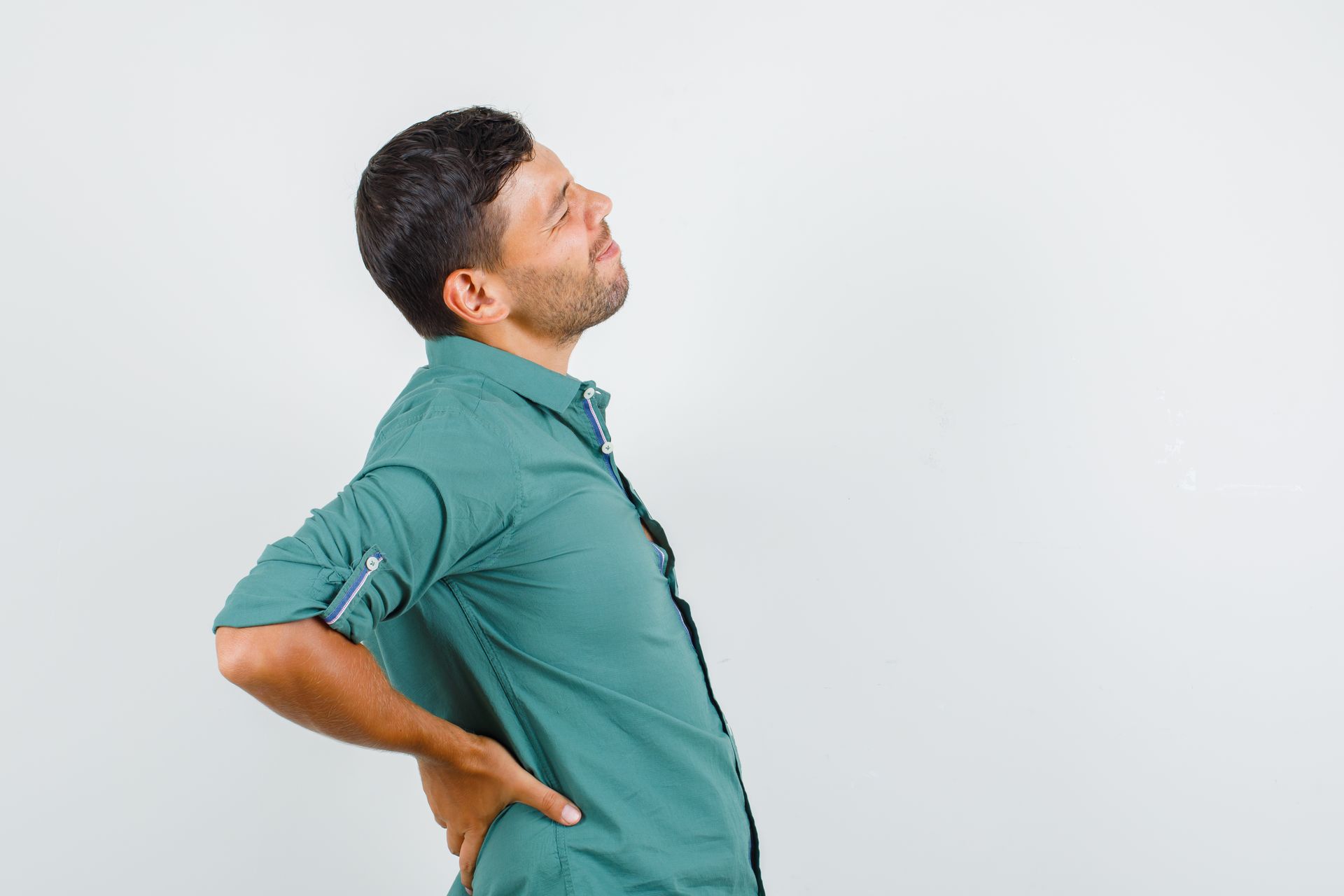 Man in a teal shirt holding his lower back and looking upward in discomfort