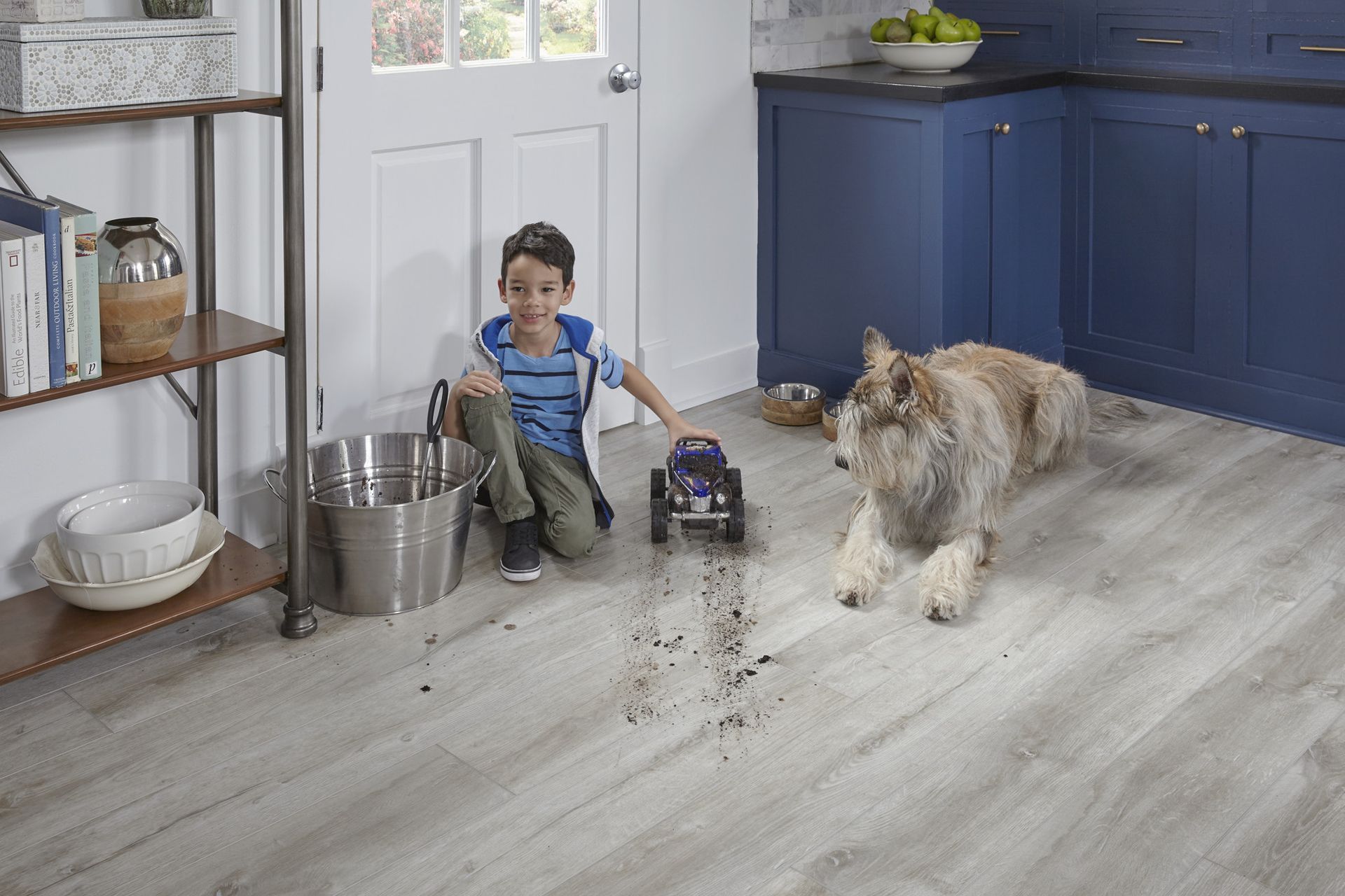 Boy and dog in kitchen with toy car and dirt on floor.