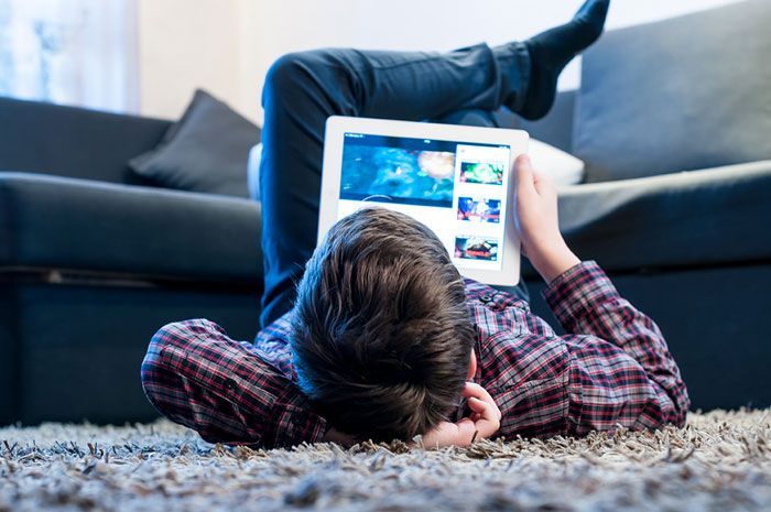 Person lying on carpet, using a tablet, feet up on couch; watching content.