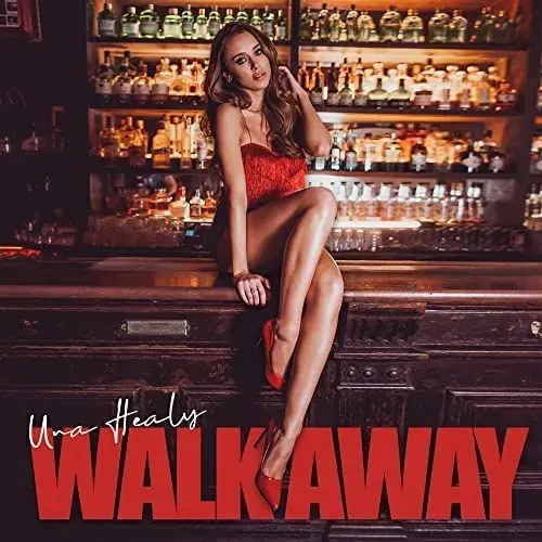 Una Healy sitting on a bar, wearing a red dress and heels. The bar is dimly lit, with bottles of liquor in the background.