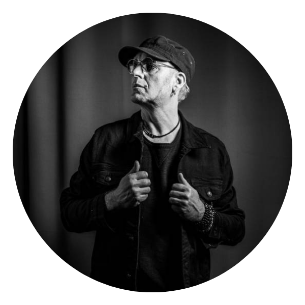 Man in black jacket and cap, looking to the side, hands on jacket, in grayscale.