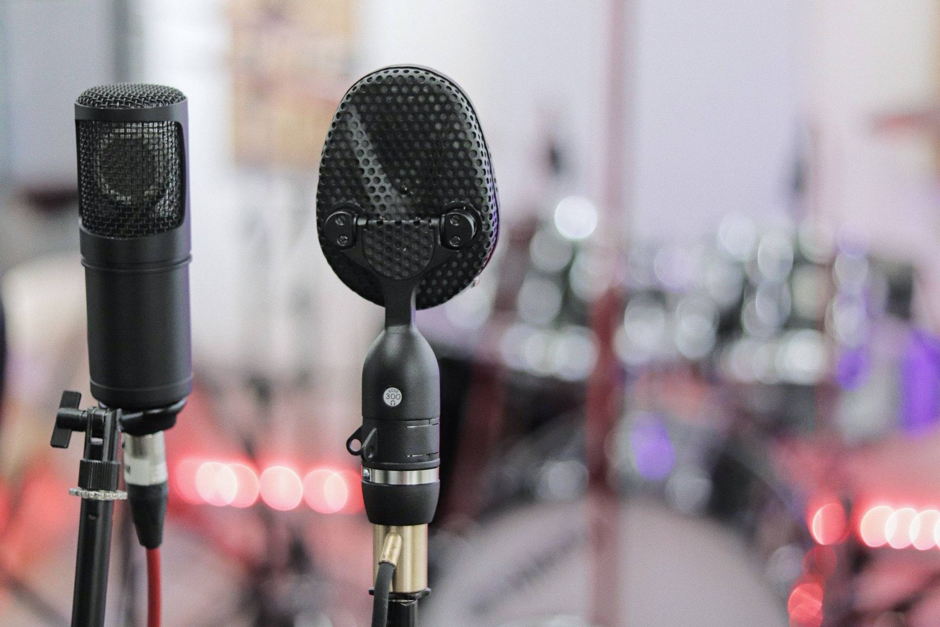 Two black microphones on stands, blurred background of a drum set and stage lights.