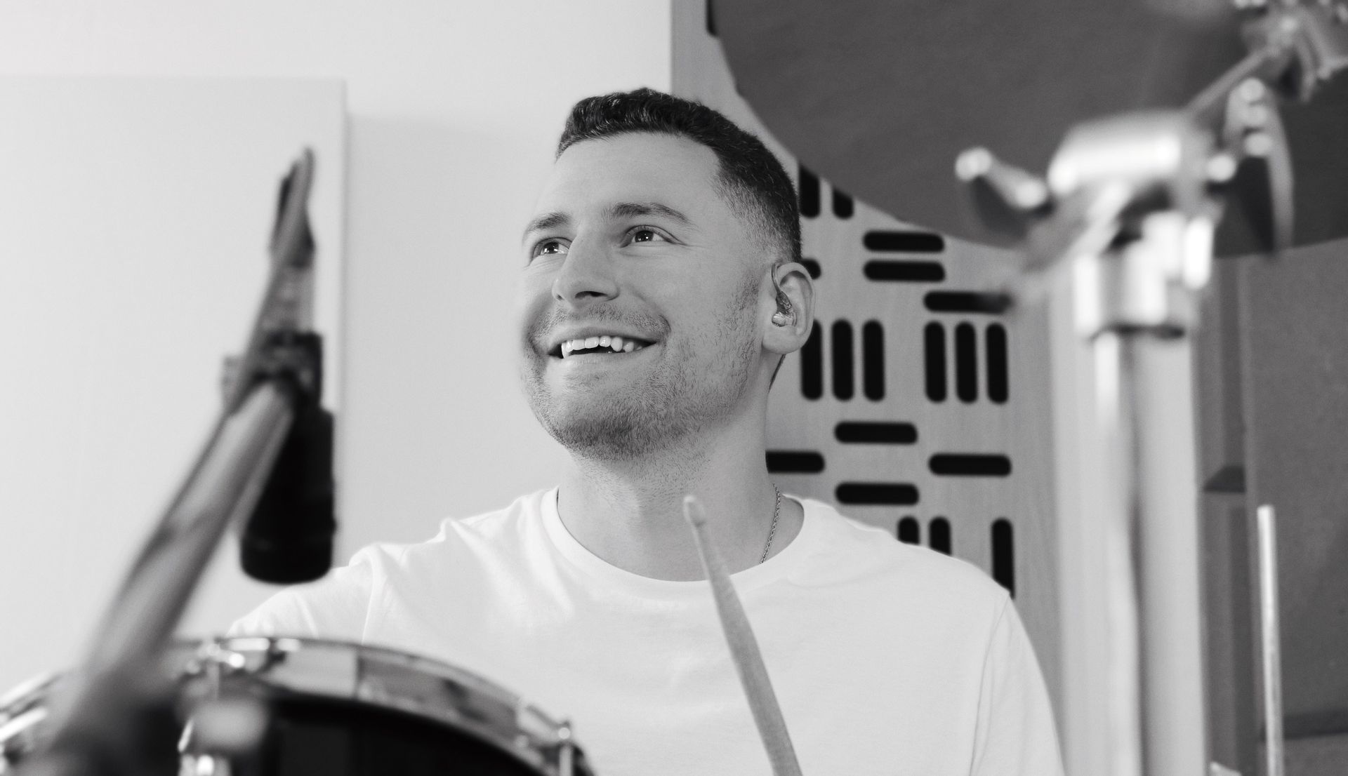 Smiling man playing drums in a studio, holding a drumstick.