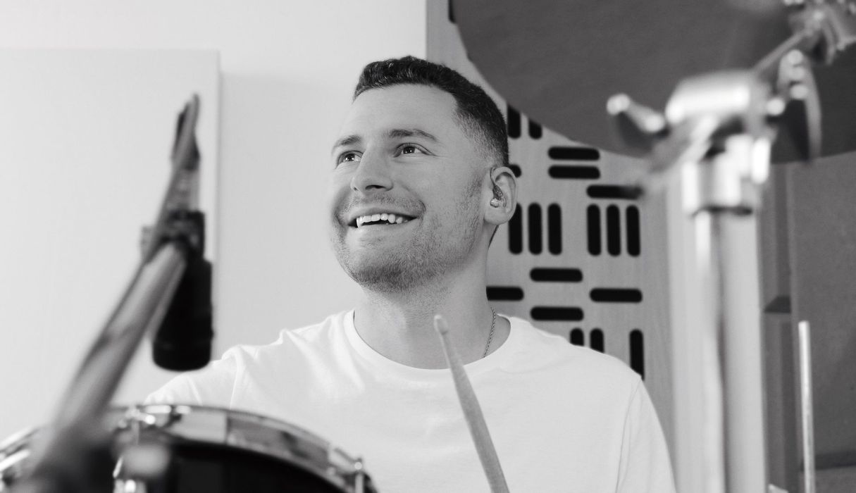 Smiling man playing drums in a studio, holding a drumstick.