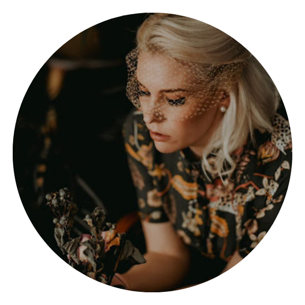Woman with blonde hair and veil looks down, holding flowers, wearing a floral top.