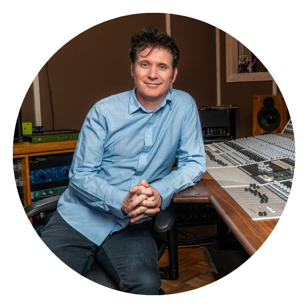 Man in blue shirt in a recording studio, leaning on a mixing console, hands clasped.