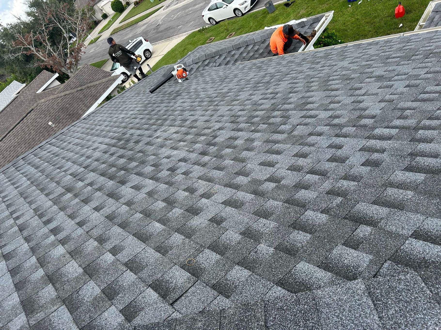 Workers installing gray asphalt shingles on a roof. One uses a leaf blower.