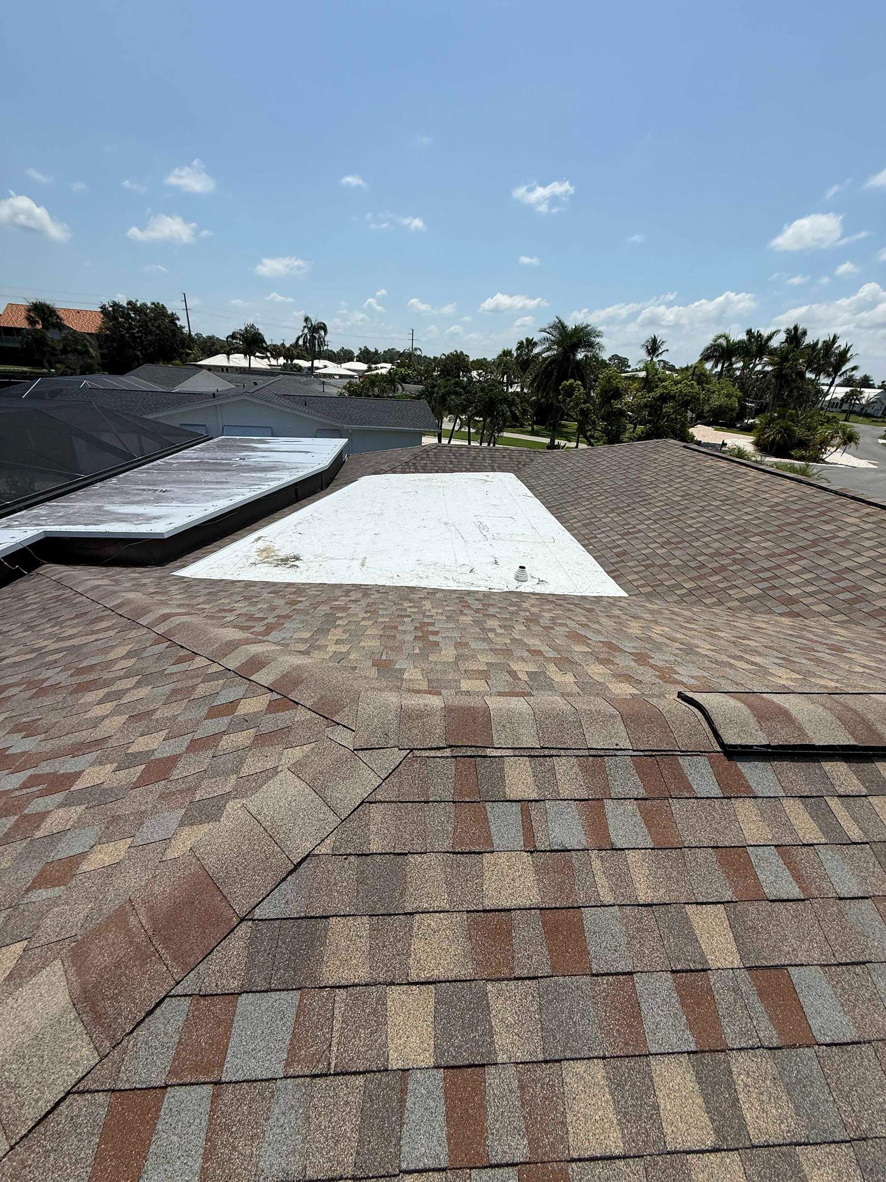 Overhead view of a roof with brown asphalt shingles and white patches against a blue sky with clouds.