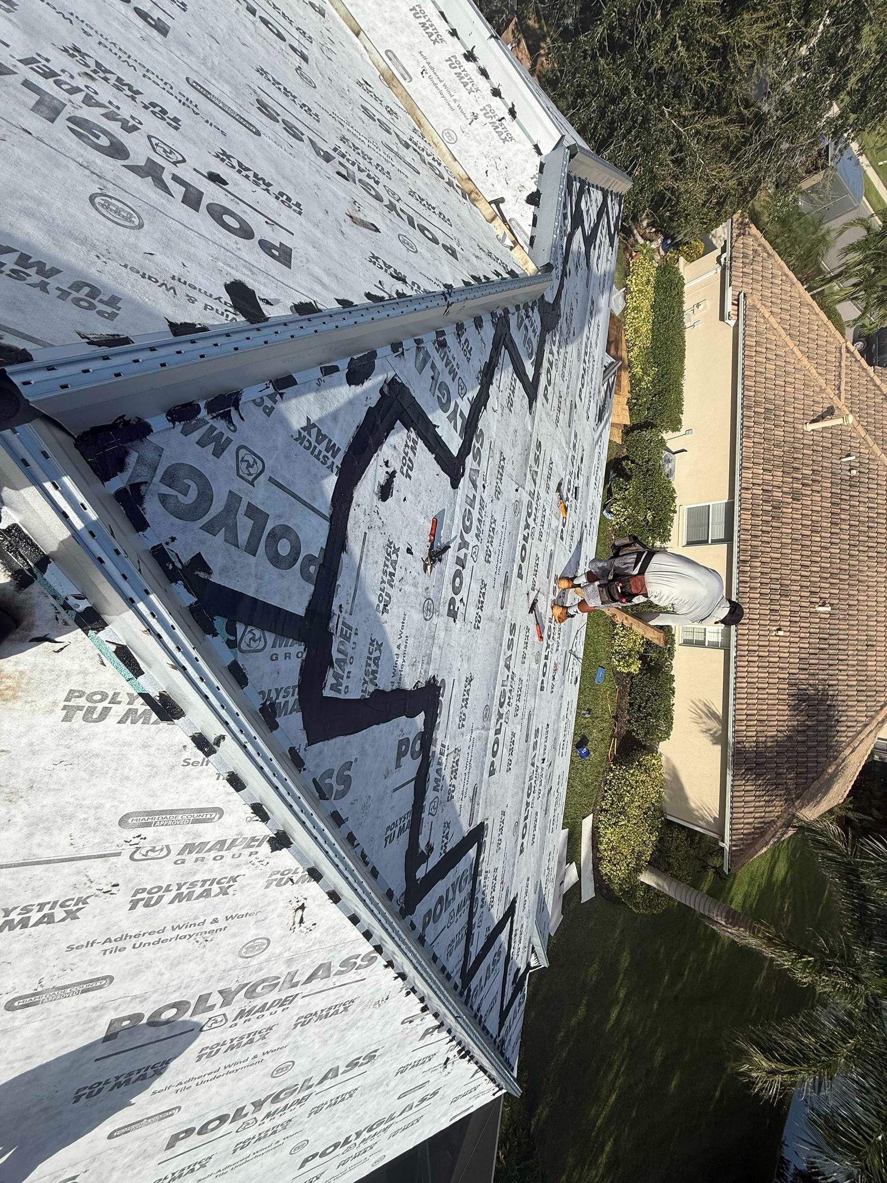 Roofer working on a roof, applying underlayment. The roof has several angles, and a house is below.