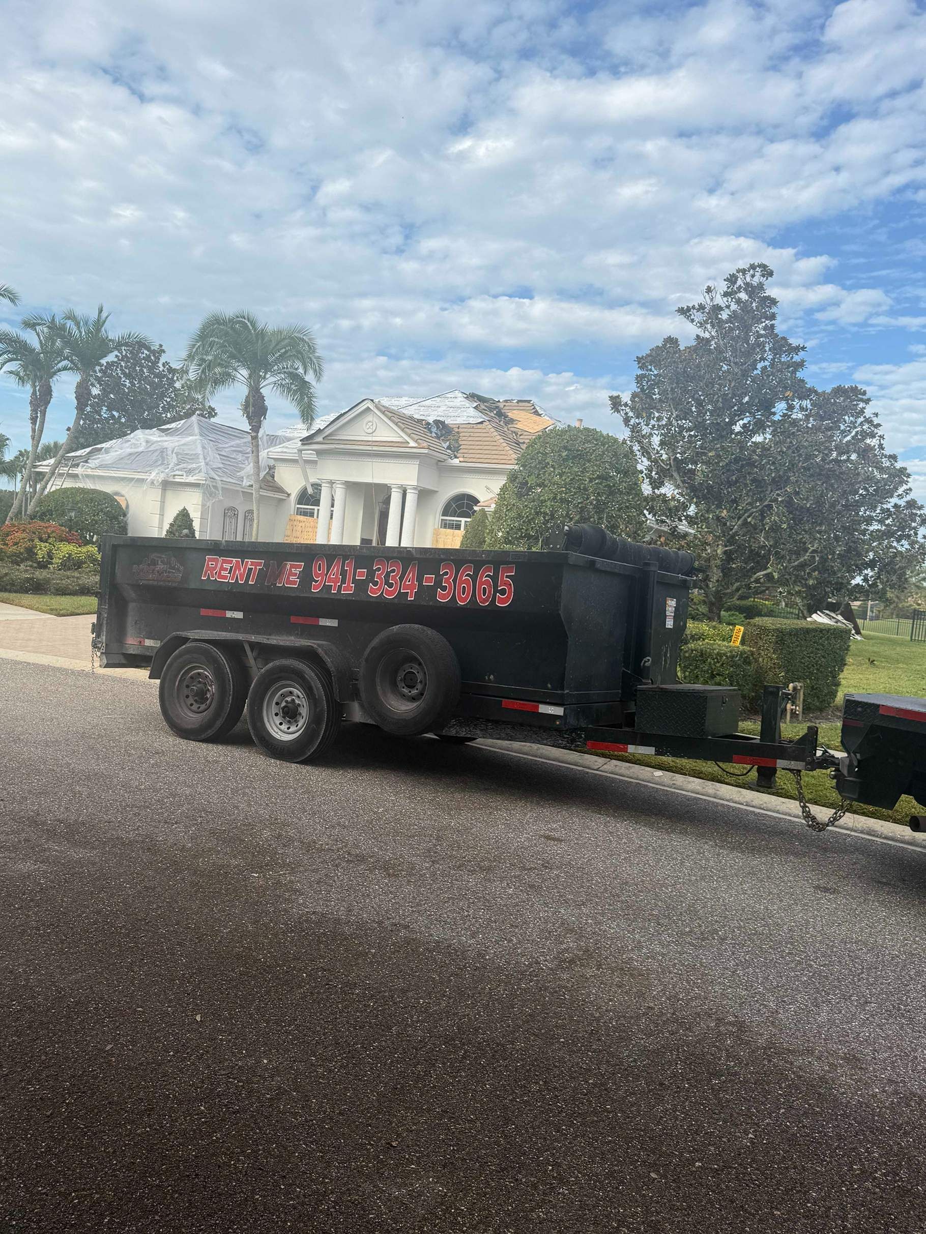 Black dump trailer on a driveway in front of a white house with palm trees, under a cloudy sky.