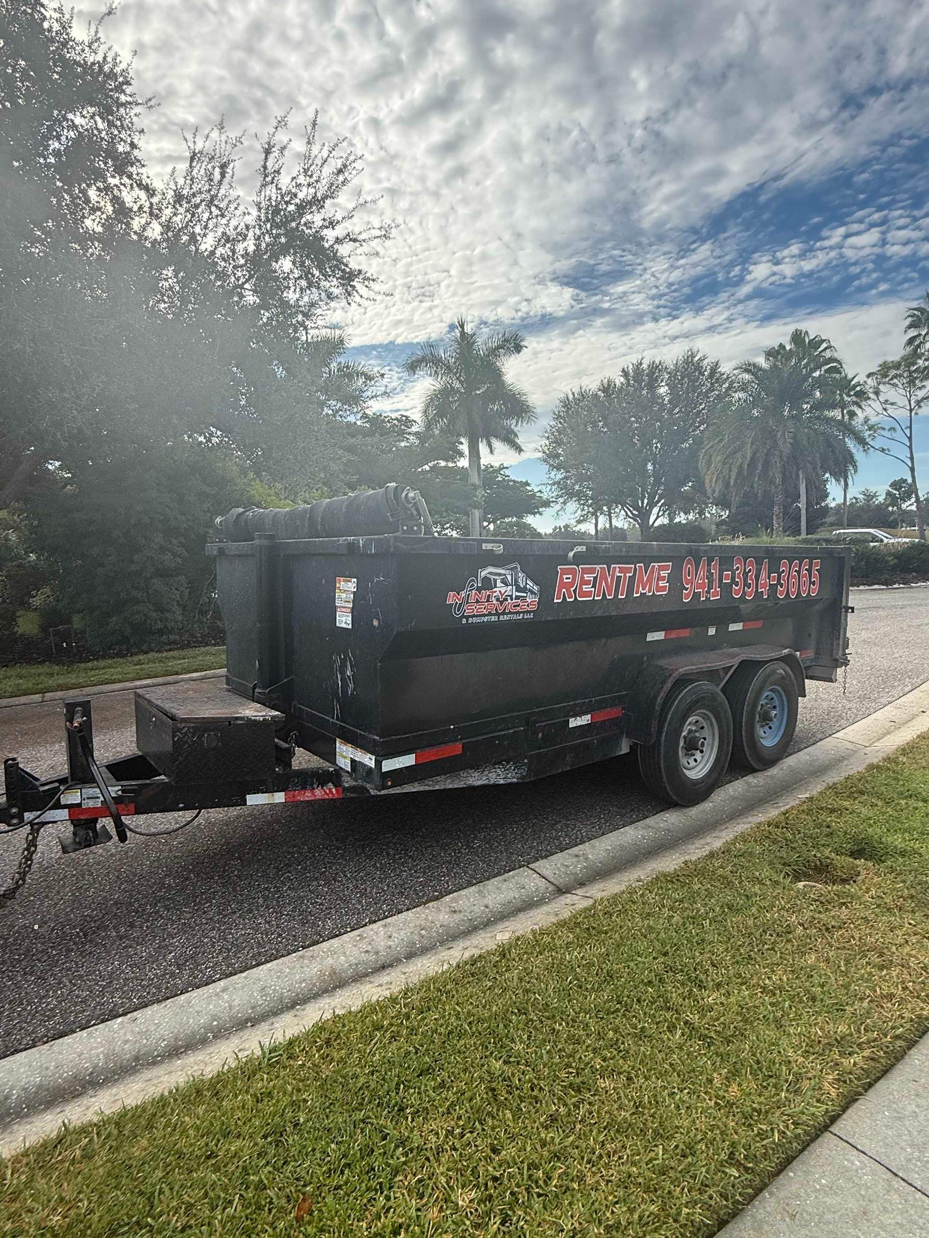 Black dumpster trailer parked on pavement near grass and trees.