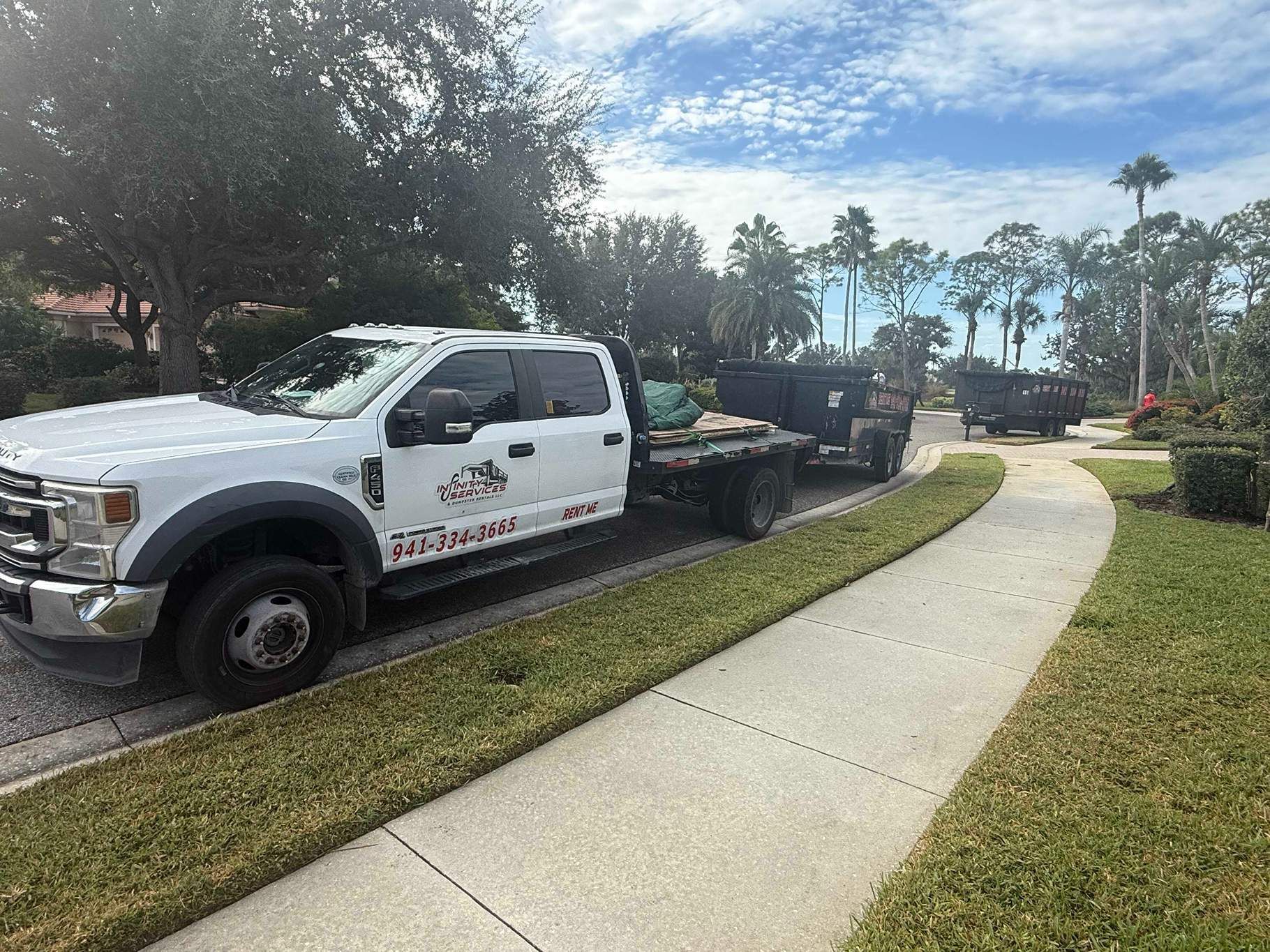White truck towing two dumpsters on a paved driveway; green grass and trees surround.