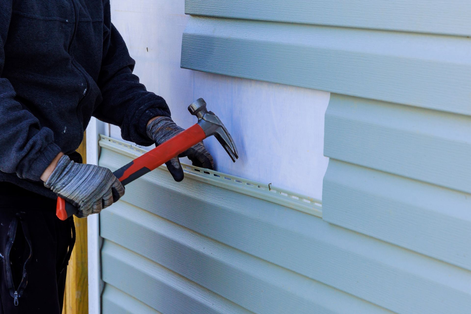 Person hammering siding on a building exterior; light blue and white color scheme.
