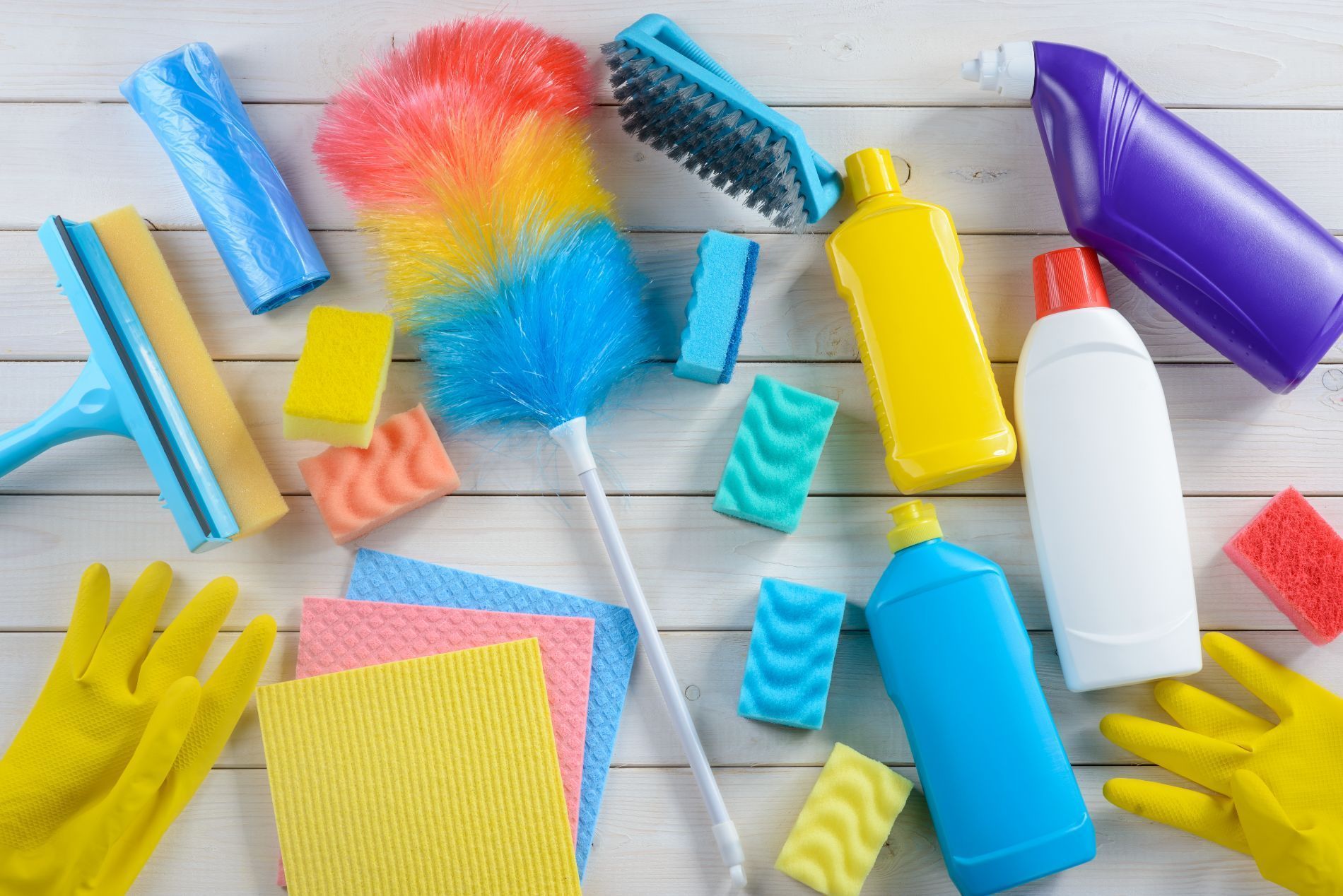 Prodotti per la pulizia disposti su una superficie di legno bianco, tra cui spugne, flaconi spray, guanti, spolverino e tergivetro.
