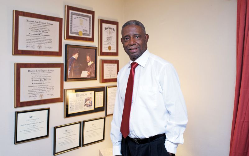 A man in a white shirt and red tie is standing in front of a wall of certificates