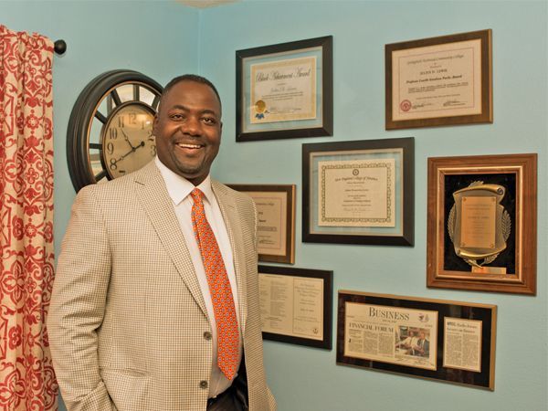 A man in a suit and tie is standing in front of a wall of framed pictures and a clock
