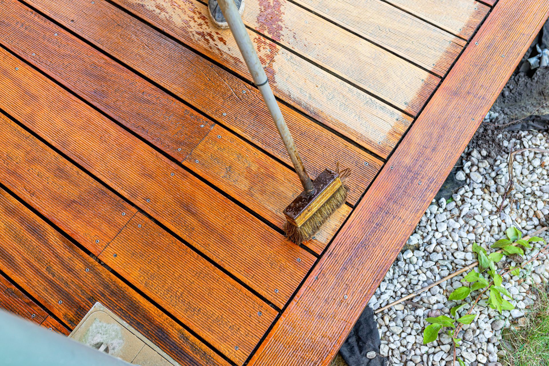 Pressure washing a wet wooden deck with orange-brown boards beside gravel and green plants