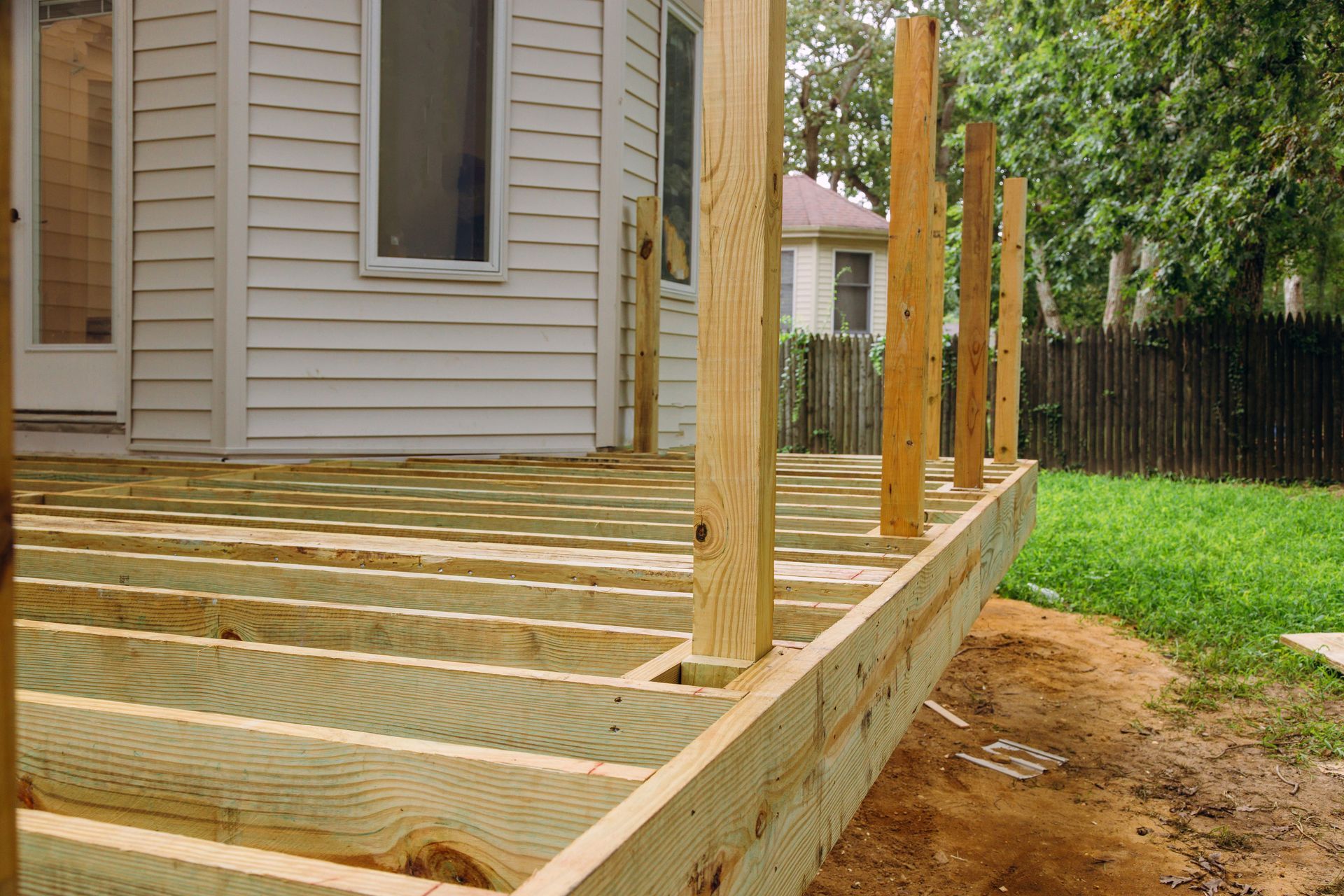 Wooden deck framing under construction beside a house, with posts and joists in a backyard.