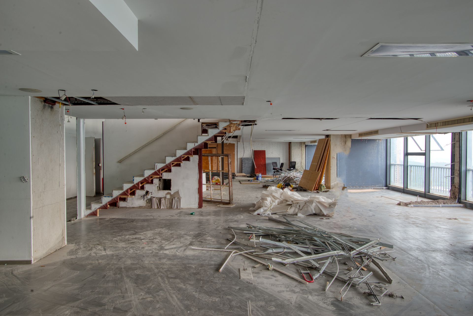 Renovation in progress in a bare interior with exposed stairs, debris piles, and large windows.