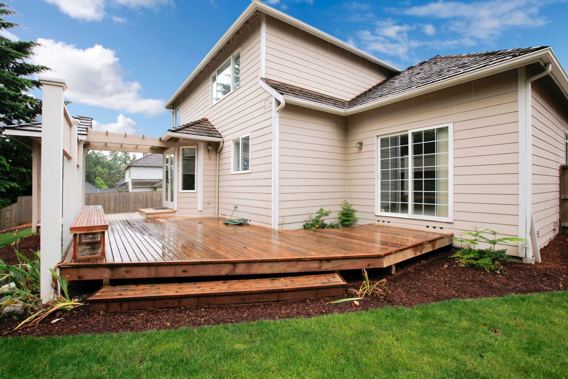 Wooden backyard deck beside a beige house with sliding glass doors and green lawn.